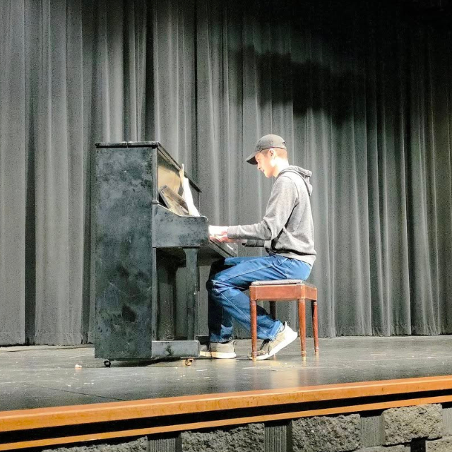 4-H Youth Member playing a black piano, he is sitting on a brown wooden stool, on a stage with black curtains
