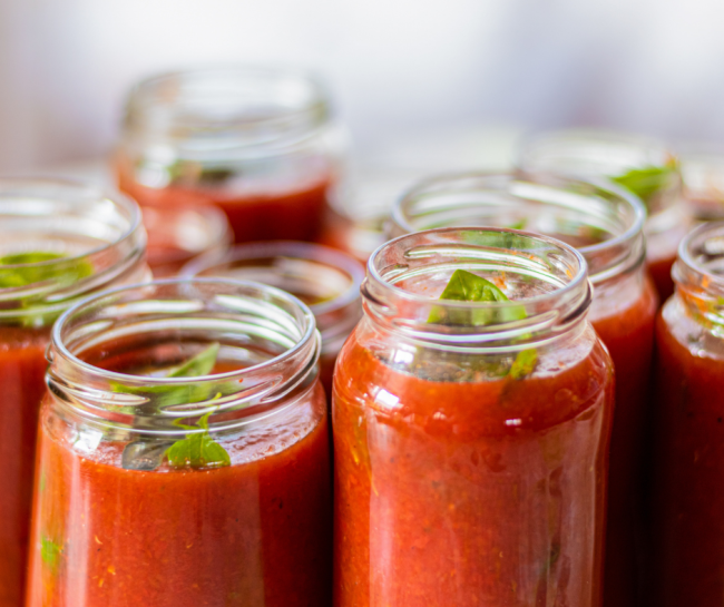 Several glass canning jars filled with red tomato sauce sit open on a countertop, ready for home food preservation. Fresh green basil leaves are visible in the jars, highlighting safe canning and food preservation practices.