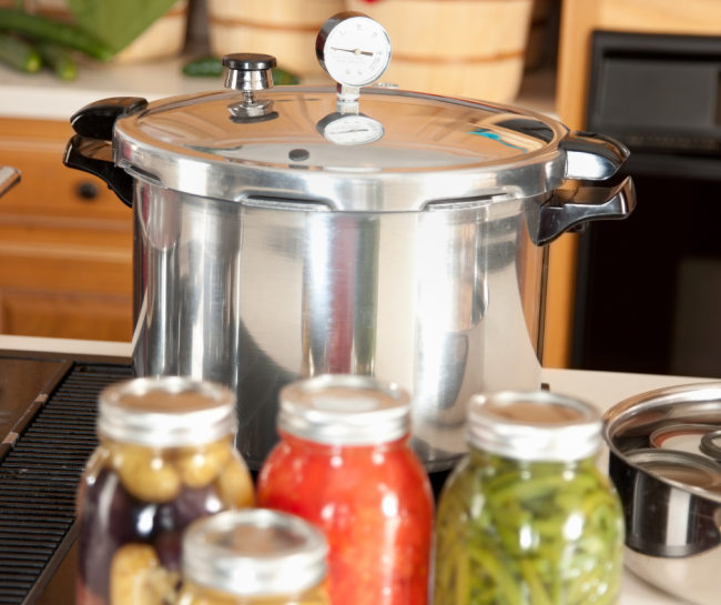 A pressure canner with a dial gauge sits on a kitchen countertop beside several sealed jars of preserved vegetables. The image represents safe home food preservation and the importance of pressure canner gauge testing for accurate canning.