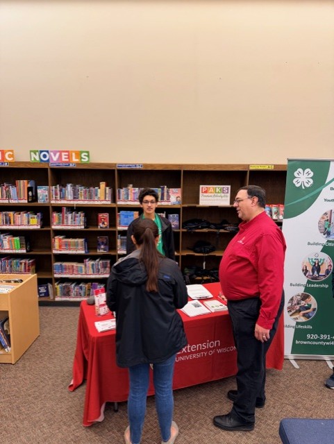 Visitors speak with staff at a UW-Madison Extension information table in a library, with bookshelves and a 4-H banner in the background.